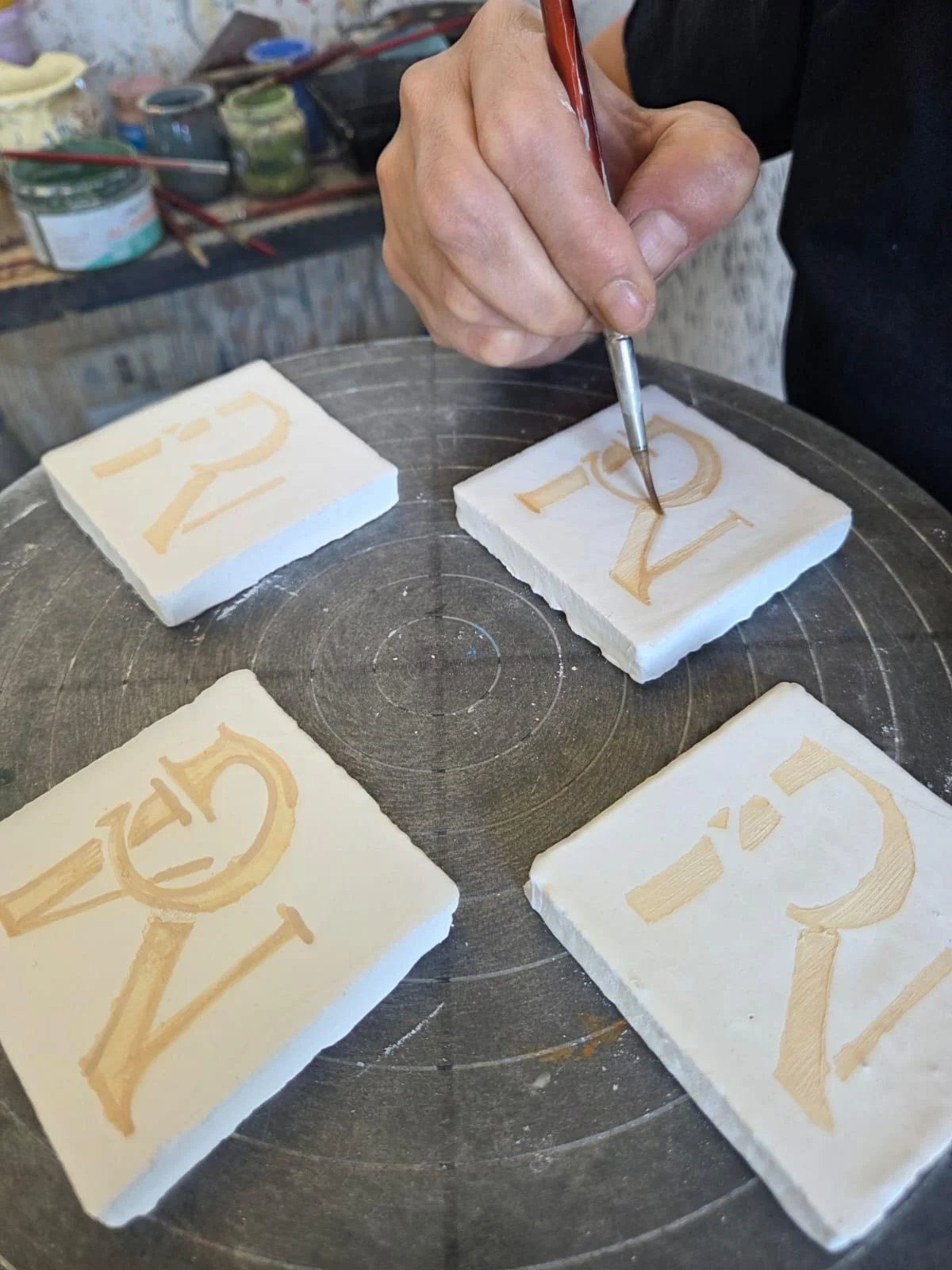 Une main peint au pinceau le monogramme MG sur des plaques de céramique blanche, posées sur un tour.
